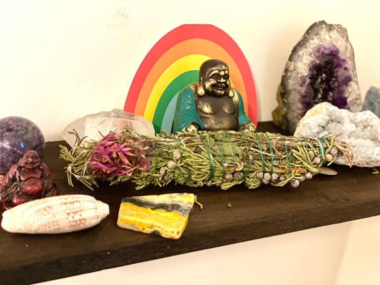 small brown shelf with herb bundle, shell, laughing Budha statue, rainbow card and colorful crystals and stones